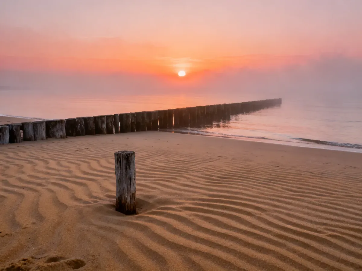 Spokojna plaża w Jantarze o wschodzie słońca z widoczną drewnianą ostrogą na tle morza i pomarańczowego nieba.